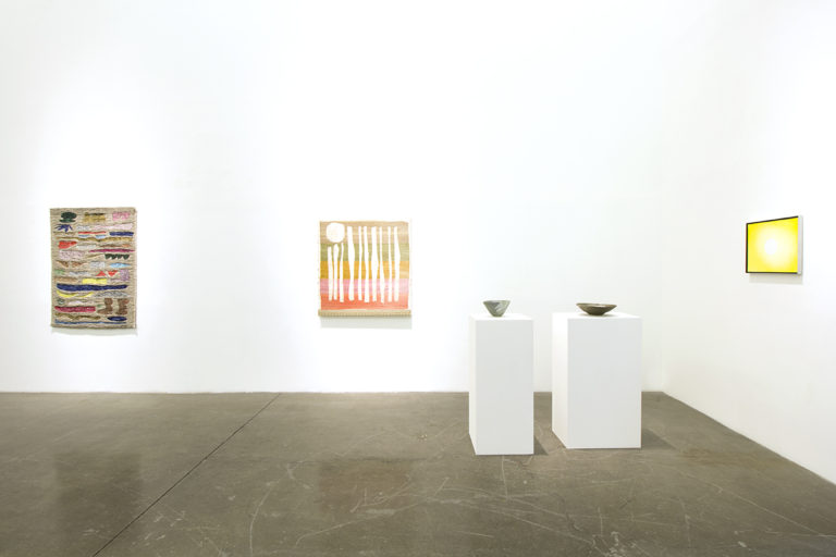 Installation image of Inclined To Blur At The Edges, of about two textile paintings and one painting on a canvas hanging on the wall of the gallery. There are two ceramic placed on a high platform