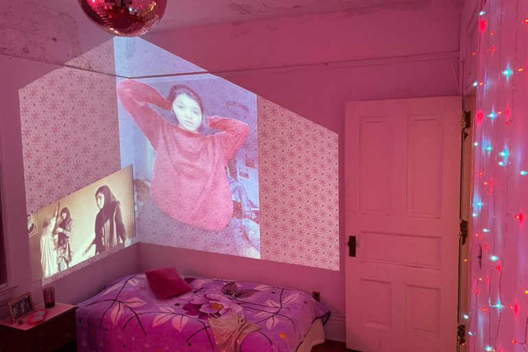 A pink teenager’s bedroom featuring gently pulsating lights and projecting on the wall is a video of women